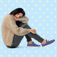 A person with chin-length dark hair sits on the floor, resting their head on their knee. They wear a beige sweater, dark pants, and Bloomscape Canvas Sneakers by My Favourite Colour is Rainbow, featuring a blue floral design. 