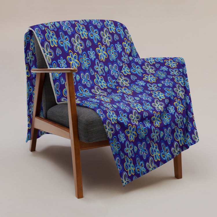 The Bloomscape Throw Blanket by My Favourite Colour is Rainbow, featuring a design of large flowers and small hearts in shades of blue and yellow on a royal blue background, is draped over a wooden armchair with grey cushions against a neutral background.