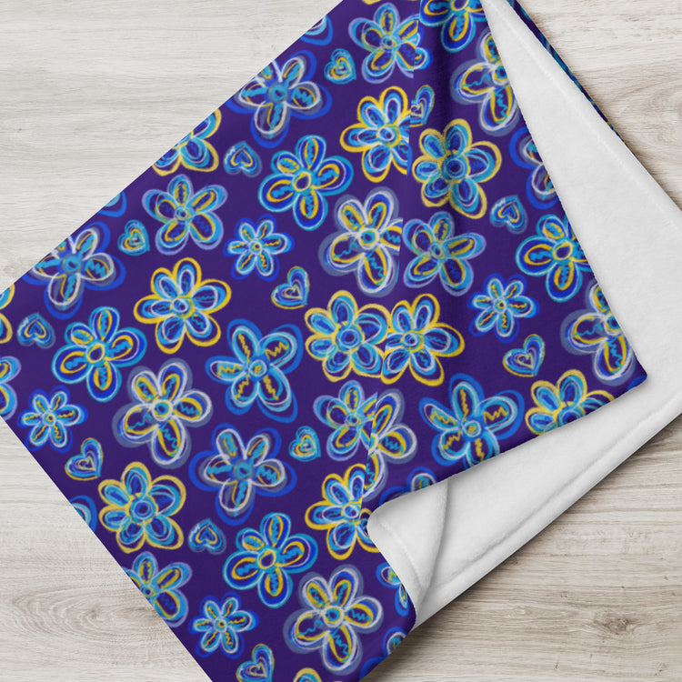 The Bloomscape Throw Blanket by My Favourite Colour is Rainbow features a blue floral and heart pattern on a royal blue background. Displayed on light wood, its folded edges reveal the white underside.