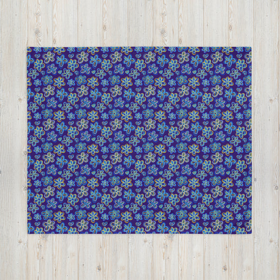 The Bloomscape Throw Blanket by My Favourite Colour is Rainbow features a design of large flowers and small hearts in shades of blue and yellow on a royal blue background. This vibrant blanket is shown on a light wooden floor.