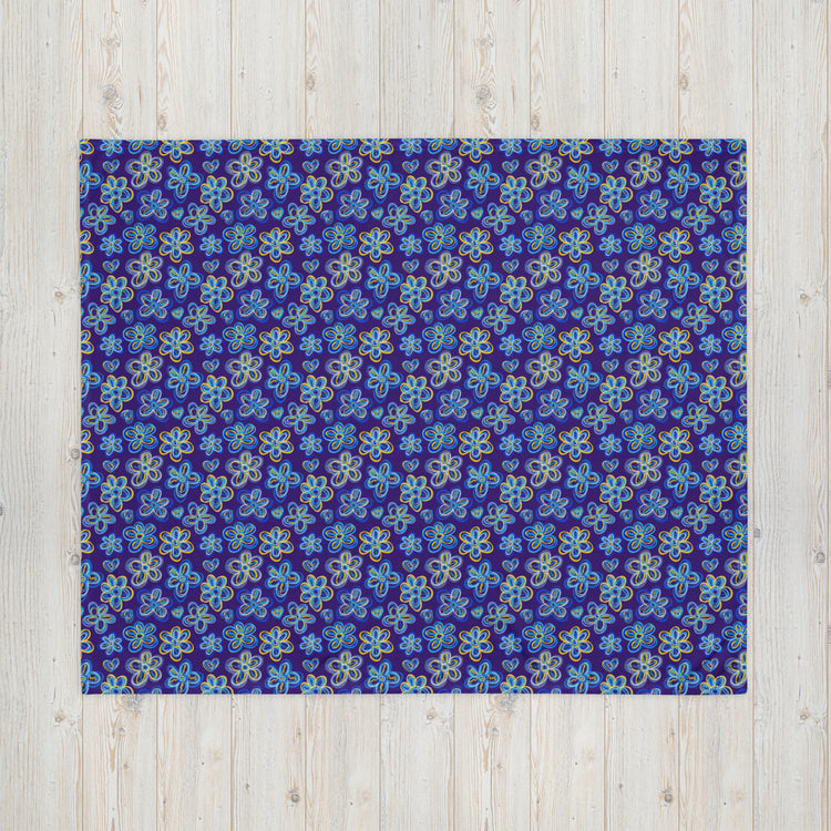 The Bloomscape Throw Blanket by My Favourite Colour is Rainbow features a design of large flowers and small hearts in shades of blue and yellow on a royal blue background. This vibrant blanket is shown on a light wooden floor.