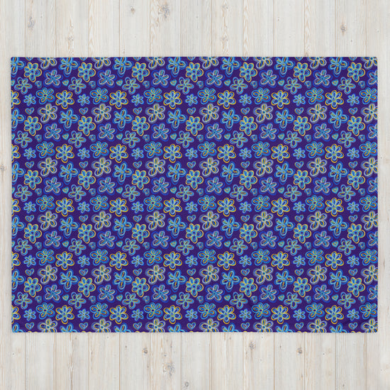 The Bloomscape Throw Blanket by My Favourite Colour is Rainbow features a design of large flowers and small hearts in shades of blue and yellow on a royal blue background. This vibrant blanket is shown on a light wooden floor.