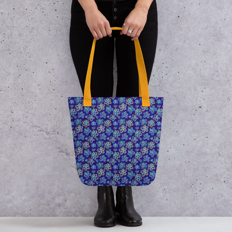 A person stands against a gray, textured wall holding the Bloomscape Tote Bag by My Favourite Colour is Rainbow. This spacious, trendy blue bag features a vibrant floral pattern with yellow handles, and they pair it with black pants and black boots.