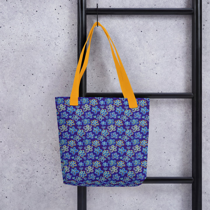 The Bloomscape Tote Bag by My Favourite Colour is Rainbow, featuring a colourful blue-and-yellow floral pattern and yellow handles, hangs on a black ladder against a gray wall.