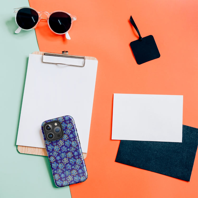 A flat lay features a clipboard with paper, sunglasses, blank cards, a black tag, and the My Favourite Colour is Rainbow Bloomscape MagSafe iPhone Case with an impact-resistant floral pattern on a split green and orange background.