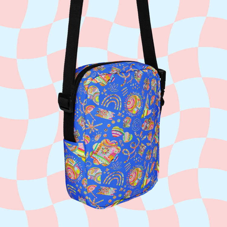 The Blue Summer Sorbet Mini Crossbody Bag by My Favourite Colour is Rainbow is a sturdy, water-resistant crossbody with adjustable black straps. It features a vibrant abstract pattern of shells and rainbows on a blue background against a pastel pink and blue checkered backdrop.