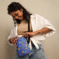 A woman with curly hair smiles while opening the Blue Summer Sorbet Mini Crossbody Bag by My Favourite Colour is Rainbow. She wears a beige top, white open shirt, light blue jeans, and displays a tattoo on her arm against a neutral background.