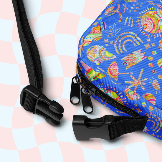 Close-up of the Blue Summer Sorbet Mini Crossbody Bag by My Favourite Colour is Rainbow, featuring abstract patterns with rainbows and stars on blue, water-resistant fabric. It has sturdy black zippers and adjustable straps with plastic buckles against a pastel pink and blue checkered backdrop.