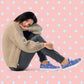 Against a pink background with white diamond patterns, a person with shoulder-length hair sits on the floor wearing a beige sweater, dark pants, and My Favourite Colour is Rainbows Blue Summer Sorbet Canvas Sneakers, resting their head on one knee.