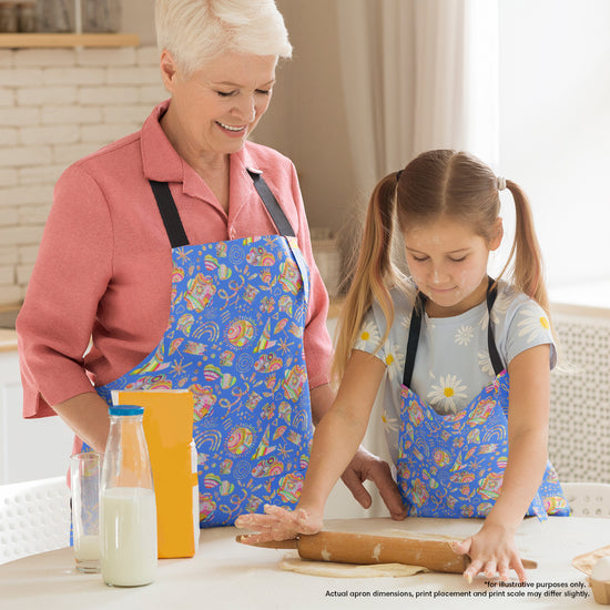 In a warmly lit kitchen, a grandmother and young girl wearing matching Blue Summer Sorbet Aprons from My Favourite Colour is Rainbow, prepare dough. Milk, flour, and an orange box sit nearby while the grandmother observes the girls focused dough rolling.