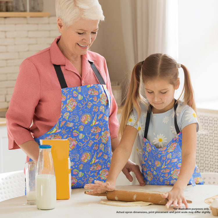 In a warmly lit kitchen, a grandmother and young girl wearing matching Blue Summer Sorbet Aprons from My Favourite Colour is Rainbow, prepare dough. Milk, flour, and an orange box sit nearby while the grandmother observes the girls focused dough rolling.