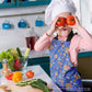 A child in a chefs hat and My Favourite Colour is Rainbow’s Blue Summer Sorbet Apron playfully holds tomatoes over their eyes, smiling. Fresh vegetables like cucumbers and bell peppers fill the kitchen counter. Utensils and a bottle appear in the vibrant background.