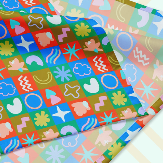 A close up of the Colour Block Bandana from My Favourite Colour is Rainbow, featuring a playful pattern of stars, waves, and spirals in blue, green, red, and yellow squares.  The design is printed on only one side of the bandana.