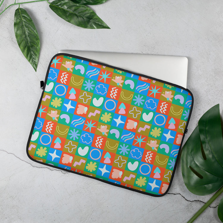 A laptop peeks out from the Colour Block Laptop Sleeve by My Favourite Colour is Rainbow, showcasing a geometric pattern in blue, red, green, and yellow. The water-resistant sleeve rests on a light gray surface, surrounded by scattered green leaves.