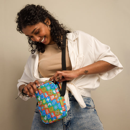 A woman with curly hair smiles while opening a Color Block Mini Crossbody Bag by My Favourite Colour is Rainbow. Shes wearing a beige top, white shirt, and jeans. The sturdy water-resistant bag hangs comfortably with its adjustable straps against the plain, neutral background.