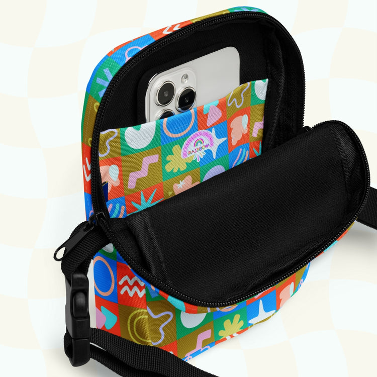 A Colour Block Mini Crossbody Bag by My Favourite Colour is Rainbow is open, displaying a smartphone inside. The sturdy bag showcases bright blue, red, and green shapes and symbols with an adjustable black strap against a light checkered pattern background.