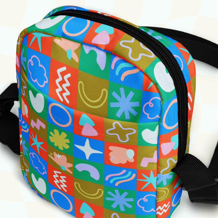 The Colour Block Mini Crossbody Bag by My Favourite Colour is Rainbow is a vibrant, small backpack adorned with abstract shapes in blue, green, pink, orange, and yellow. It has a black zipper and sturdy black straps against a textured backdrop. This water-resistant bag suits any adventure.