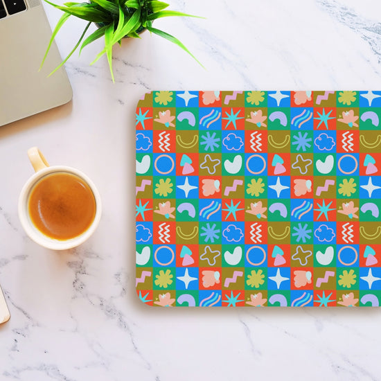 A laptop and coffee cup sit on a white marble surface beside a Colour Block Mouse Pad from My Favourite Colour is Rainbow featuring abstract shapes in blue, red, green, and beige. A small green plant decorates the top left corner.