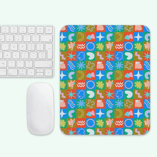 The Colour Block Mouse Pad by My Favourite Colour is Rainbow, featuring a grid of abstract patterns and shapes with squares showing circles, stars, waves, and flowers in blue, green, red, and orange tones, sits next to a white keyboard and mouse.