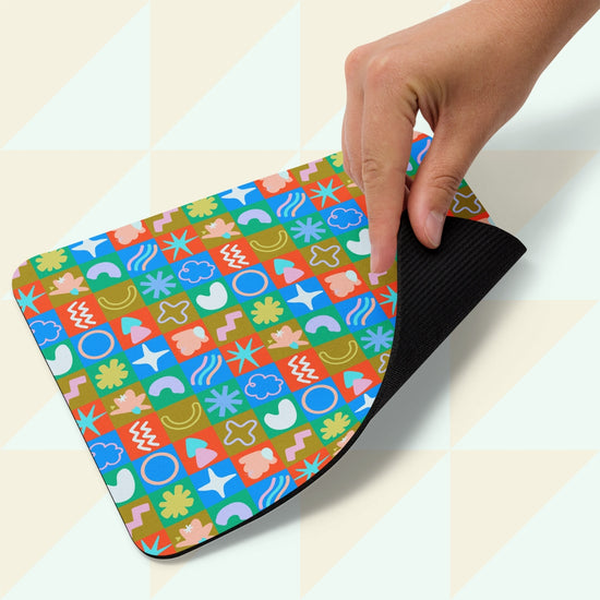 A hand lifts the corner of the Colour Block Mouse Pad, designed by My Favourite Colour is Rainbow, featuring a geometric pattern with circles, squares, and wavy lines in bright colors on top and a black underside. It rests on a light checkered background.