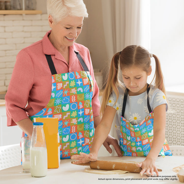 In a bright kitchen, a grandmother and granddaughter bake together, both wearing Colour Block Aprons by My Favourite Colour is Rainbow. As the girl rolls dough with her grandmother smiling nearby, milk and cereal on the table enhance the vibrant setting.