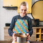 In a kitchen with a yellow cupboard and various appliances, a woman wearing the My Favourite Colour is Rainbows Colour Block Apron holds a springform pan with a cake. She smiles, focused on her task, as her vibrant attire complements the kitchen perfectly.