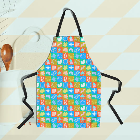 The Colour Block Apron by My Favourite Colour is Rainbow features a vibrant design of abstract shapes within a checkerboard pattern, in red, green, yellow, and blue. A white container holding cooking utensils and a chopping board are visible in the background.