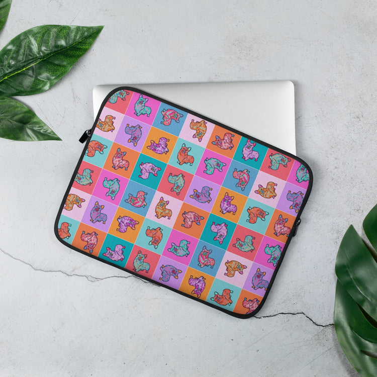 The 13" Crazi Corgis Laptop Sleeve by My Favourite Colour is Rainbow features a vibrant pop art-style design of illustrated corgis on a checkered background in shades of pink, red, orange, blue and teal. A laptop partially peeks out from this case, which rests on a concrete surface with palm leaves.