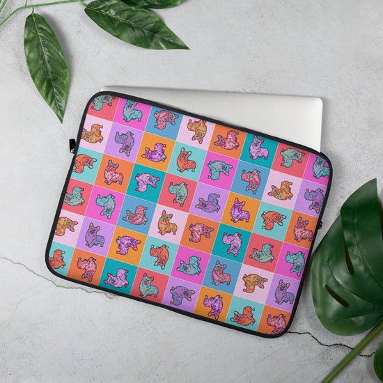 The 15" Crazi Corgis Laptop Sleeve by My Favourite Colour is Rainbow features a vibrant pop art-style design of illustrated corgis on a checkered background in shades of pink, red, orange, blue and teal. A laptop partially peeks out from this case, which rests on a concrete surface with palm leaves.