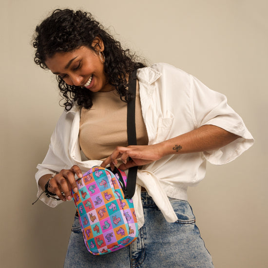 A person with curly hair in a beige top, white shirt, and jeans smiles while adjusting the colorful Crazi Corgis Mini Crossbody Bag by My Favourite Colour is Rainbow against a plain beige background.