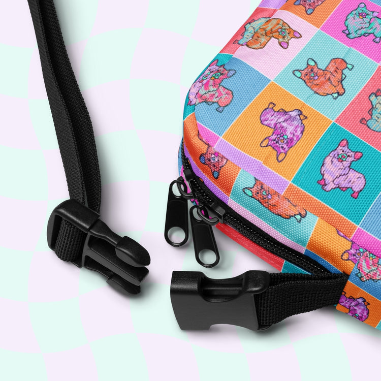 A close-up of the Crazi Corgis Mini Crossbody Bag from My Favourite Colour is Rainbow, showcasing a colorful pattern of cartoon sheep in pink, orange, and blue squares. The water-resistant bag features black zippers and adjustable straps with clips against a pastel pink and green checkered background.