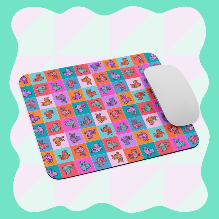 The Crazi Corgi Mouse Pad by My Favourite Colour is Rainbow features cartoon dogs in various poses with a checkerboard background of pink, teal, red, and purple squares. A white mouse rests on it against an aqua backdrop with a wavy pattern.