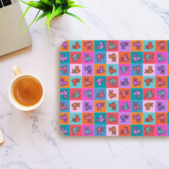 The Crazi Corgi Mouse Pad by My Favourite Colour is Rainbow showcases cartoon dog heads on a marble surface, with a coffee cup and part of a silver laptop beside it, plus a small plant nearby.