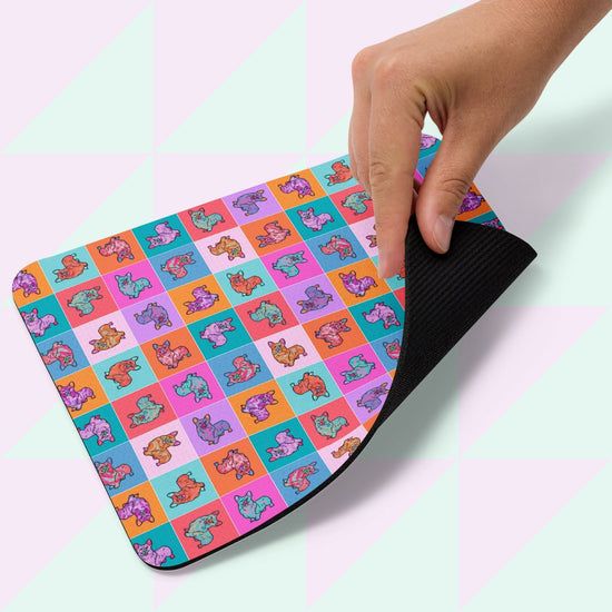 A hand lifts a Crazi Corgi Mouse Pad by My Favourite Colour is Rainbow, featuring a grid of colorful cartoon dinosaur illustrations. The underside is black against a pastel geometric background.