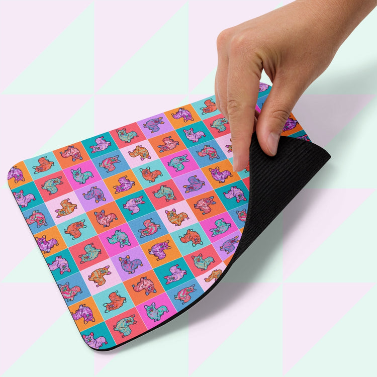 A hand lifts a Crazi Corgi Mouse Pad by My Favourite Colour is Rainbow, featuring a grid of colorful cartoon dinosaur illustrations. The underside is black against a pastel geometric background.