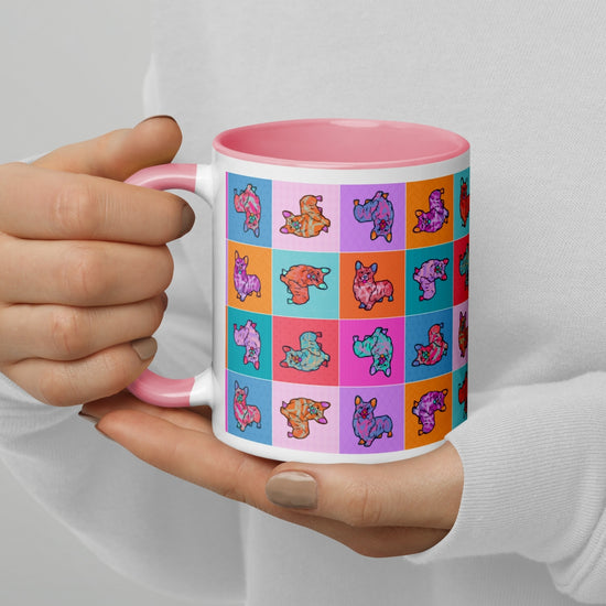 A coffee enthusiast holds the Crazi Corgis Mug by My Favourite Colour is Rainbow, a durable ceramic mug with a white exterior and pink interior. It features colorful, patterned animal illustrations including adorable corgis on a vibrant, playful grid with varying background colors.