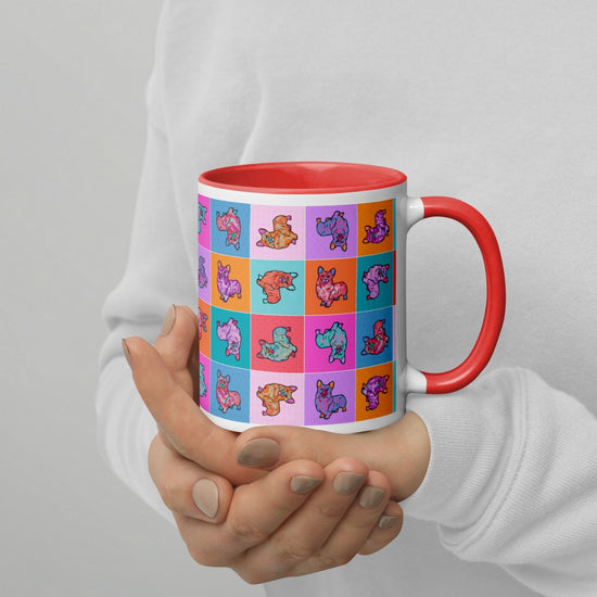 A coffee enthusiast holds a Crazi Corgis Mug by My Favourite Colour is Rainbow, featuring a red handle and durable ceramic. This charming mug showcases a vibrant square pattern with cartoon-style dinosaurs. The person wears a cozy white sweater.