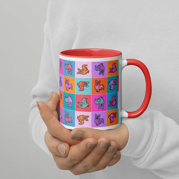 A coffee enthusiast holds a Crazi Corgis Mug by My Favourite Colour is Rainbow, featuring a red handle and durable ceramic. This charming mug showcases a vibrant square pattern with cartoon-style dinosaurs. The person wears a cozy white sweater.