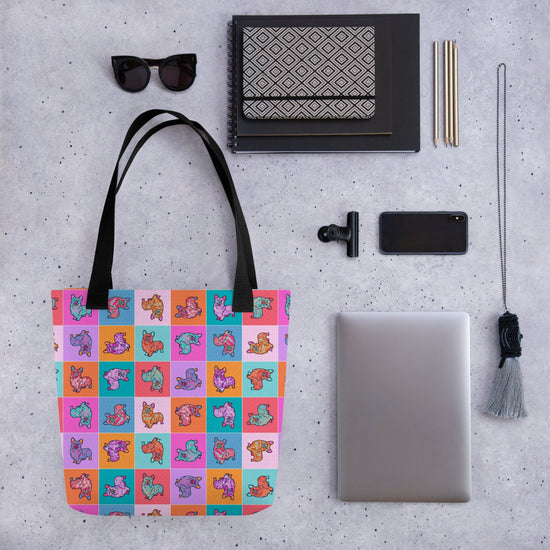 Flat lay featuring a Crazi Corgis Tote Bag by My Favourite Colour is Rainbow, sunglasses, notebook, black phone, laptop, tassel keychain, and pens on a speckled gray surface.