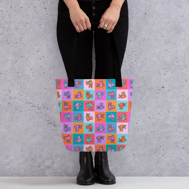 A person in black pants and boots holds a trendy Crazi Corgis Tote Bag by My Favourite Colour is Rainbow, boasting ample space with a pop art corgi pattern on a checkerboard of pink, turquoise, orange, and purple squares.