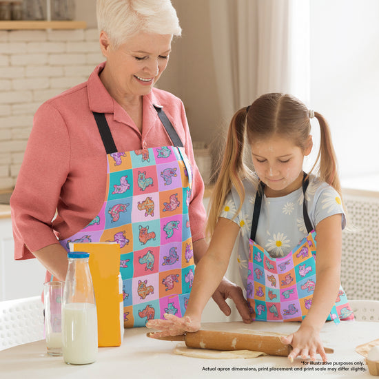 In the kitchen, a grandmother and granddaughter wear Crazi Corgis Aprons from My Favourite Colour is Rainbow. The grandmother smiles as her granddaughter rolls dough with a rolling pin. Milk and juice cartons on the table enhance their cosy scene.