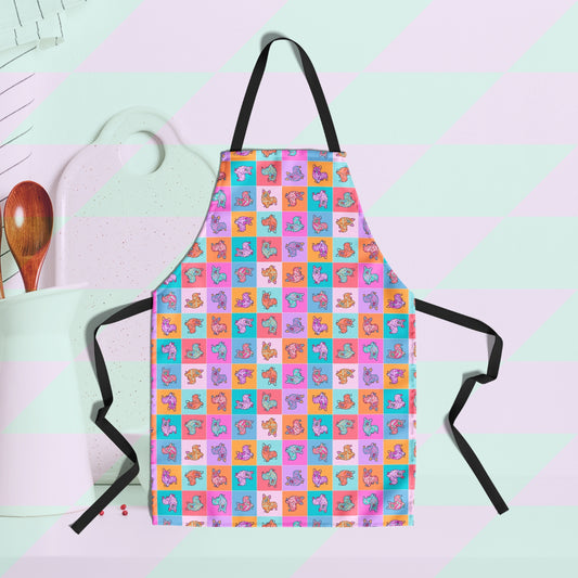 A Crazi Corgis apron by My Favourite Colour is Rainbow, featuring a checkerboard design of colourful pop-art corgis in blue, orange, purple, pink and red.  The background features a wooden spoon and a white chopping board.