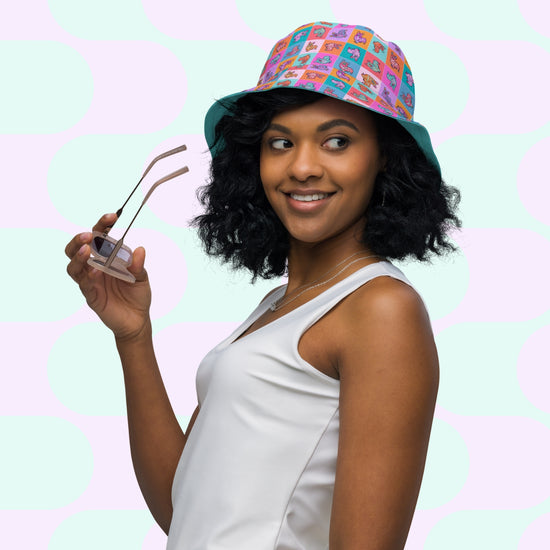 A woman with curly hair smiles, holding pink sunglasses and wearing the Crazi Corgis Reversible Bucket Hat by My Favourite Colour is Rainbow and a white sleeveless top. A pastel polka dot background adds a playful touch to her stylish streetwear look.