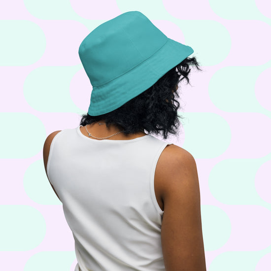 A person with curly hair wears the Crazi Corgis Reversible Bucket Hat by My Favourite Colour is Rainbow, with a white sleeveless top, standing against a pastel green and pink geometric pattern backdrop that makes this streetwear accessory pop.