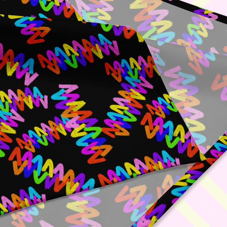 A closeup of the Electric Zigzag Bandana by My Favourite Colour is Rainbow, featuring vibrant red, yellow, blue, pink, and green zigzags on a black backdrop.  The design is printed on only one side of the bandana.