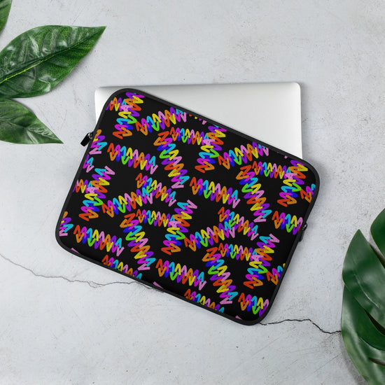 A 13" Electric Zigzag laptop sleeve from My Favourite Colour is Rainbow, featuring vibrant red, blue, purple, and orange patterns, sits on a light gray surface with green leaves along the edges. The sleeve partially covers a laptop.