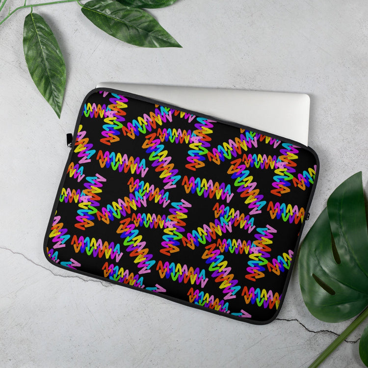 A 15" Electric Zigzag laptop sleeve from My Favourite Colour is Rainbow, featuring vibrant red, blue, purple, and orange patterns, sits on a light gray surface with green leaves along the edges. The sleeve partially covers a laptop.