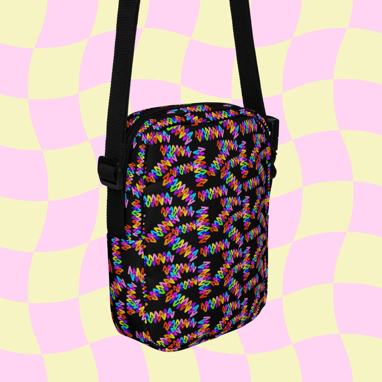 The Electric Zigzag Mini Crossbody Bag by My Favourite Colour is Rainbow features a vibrant design with a wavy pink and yellow checkerboard on a black backdrop. The adjustable black strap complements the striking pattern perfectly.