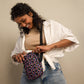 A woman with curly hair, in a beige top, white open shirt, and blue jeans against a plain background, smiles while opening the My Favourite Colour is Rainbow Electric Zigzag Mini Crossbody Bag. Its adjustable straps enhance her vibrant look with added versatility.