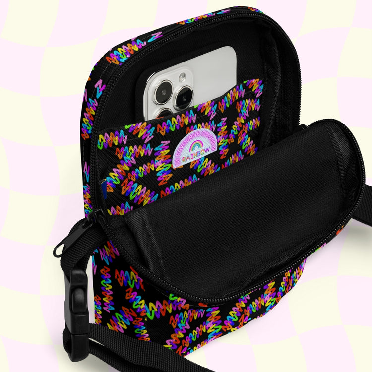 A black Electric Zigzag Mini Crossbody Bag by My Favourite Colour is Rainbow is open, showing a smartphone inside. Attached to its outer pocket is a small round badge with text and a rainbow. The background has a pastel light checkered pattern.